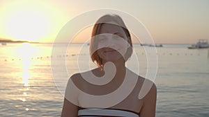 30s woman in swimsuit walking to the sea, turning to camera and smiling. Female enjoys dawn on the beach. Back view of