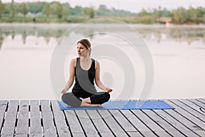 A 36-year-old young Caucasian woman practices yoga in the lotus position outdoors near the river on a wooden pier in the morning.