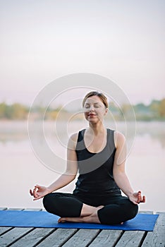 A 36-year-old young Caucasian woman practices yoga in the lotus position outdoors near the river on a wooden pier in the morning.