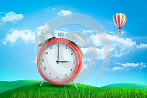 3d rendering of red retro alarm clock showing 3 o`clock standing on green grass under blue sky with hot air balloon in