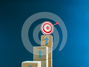 3d target icon on the top of wooden cube blocks, bar graph chart steps with up arrows on blue background.