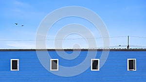 4 Glass windows on blue wooden wall of row house with birds perching on electric cable lines against blue sky