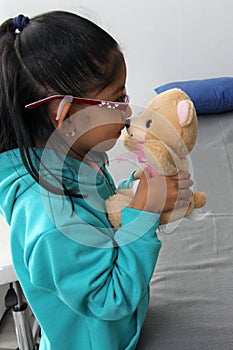 4-year-old brunette Latina girl with glasses represents mistreatment and physical abuse in her teddy bear with bandages on her hea