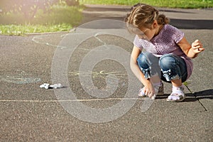 4 year old girl in casual clothes sits on the pavement and draws a line with chalk on the street