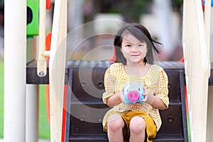 4-year-old girl uses two hands to hold piggy bank shaped blue and pink nose. Asian girl is sitting at player staircase. Happy with