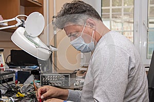 A 40-45-year-old man in a medical mask checks the electrical Board from the computer with a tester, close-up, Repair of computer e