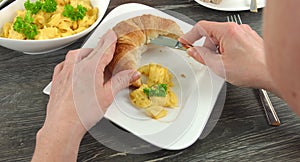 4K: Cut open croissant and some scrambled eggs lie on an angular white plate. A woman is putting some fresh scrambled eggs to the