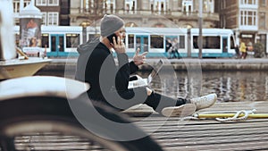 4K European businessman working outside. Man talking on the phone sitting with laptop. River boat and tram passing by.