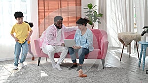 4K Father and Mother sit on couch using laptop with children running around