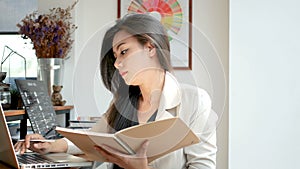 4K footage, beautiful Asian business woman sitting with computer laptop keying data, reading and flip over note book page