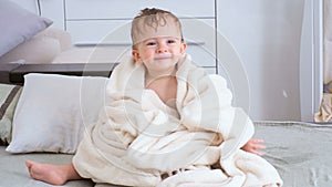 4K footage of cute smiling baby boy covered in towel sitting on bed after bathing