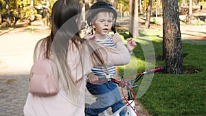 4k footage of little toddler boy sitting on bicycle at park while his caring mother putting on protective helmet before