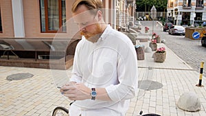 4k footage of stylish red bearded man in white shirt sitting on vintage bicycle and typing message on his mobile phone