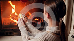 4k footage of young woman in sweater warming and rubbing her hands at burning fireplace in house