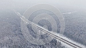 4K. Road in the winter forest with driving cars at snowfall. Aerial panoramic view.