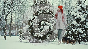 4k slow motion beautiful smiling young woman in parka, knitted red beanie and mittens walks with her Welsh Corgi dog