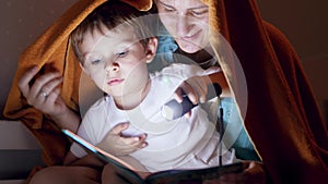 4k video of happy smiling mother with little son sitting under blanket at night and reading book with flashlight