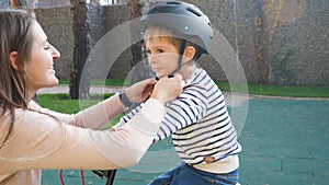 4k video of happy smiling young mother putting protective helmet on her little son before riding bicycle at park