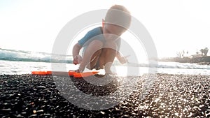 4k video of little 3 years old boy collecting stones and seasheels on sea beach on beautiful sunset