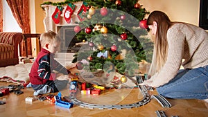 4k video of little boy playing with his mother with toy train and railroad under Christmas tree at living room. Child
