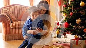 4k video of smiling young mother with her little boy watching cartoons on mobile phone under Christmas tree