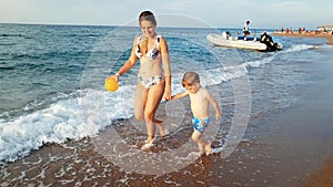 4k video of young mother holding her child by hand walking on the sandy sea beach. Toddler boy relaxing on beach with