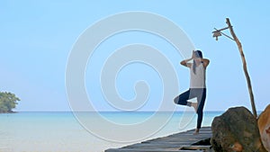 4K. A woman standing on one leg while practicing yoga on wooden bridge over the sea during summer vacation. Attractive young asian