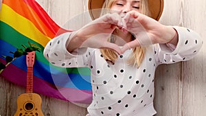 4k Young blonde millennial hippie woman showing heart sign with hands with rainbow LGBTQ flag at home balcony. Peace and