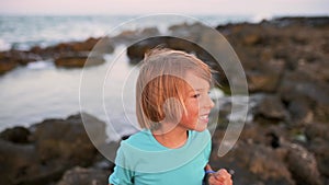 A 5-year-old boy with long hair enthusiastically shares something while standing by the sea in the evening.