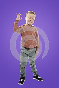 A 5-year-old boy stands and shows the ok sign with his hand. A child in jeans and a brown sweater. Success and positive. Purple