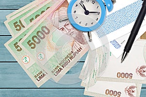 5000 Indonesian rupiah bills and alarm clock with pen and envelopes. Tax season concept, payment deadline for credit or loan.