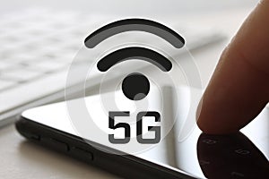 5G network concept with finger touching smartphone with screen and keybord in background. Wireless internet symbol in front of dis