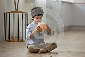 5s Indian boy opening pink piggy bank, sitting on floor