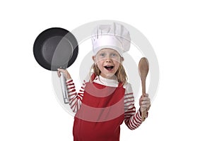 6 or 7 years old little girl in cooking hat and red apron playing cook smiling happy holding pan and spoon
