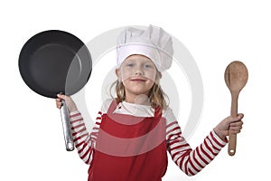 6 or 7 years old little girl in cooking hat and red apron playing cook smiling happy holding pan and spoon