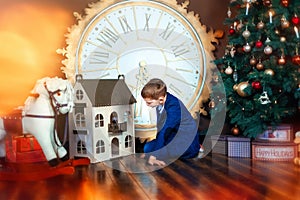 6 years old boy sit on floor and look in dollhouse. Rocking horse and big clock near. Christmas eves with beautiful christmas tree