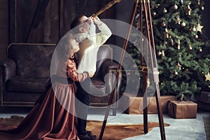 6 years old boy in vintage clothes and curly girl in retro brown dress looking in telescope with christmas tree on background.