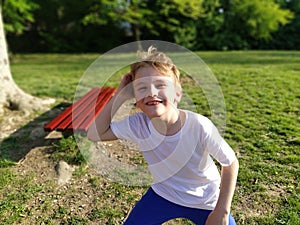 A 7-year-old boy with blond hair in a white T-shirt and blue pants poses in a city park. Child fooling around, squatting and