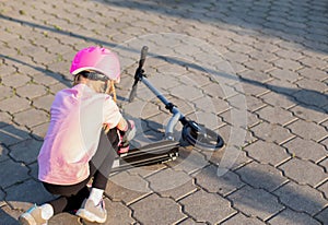 A 7-year-old girl in a pink helmet and protective gear fell off a scooter. The concept of safe riding a scooter and a