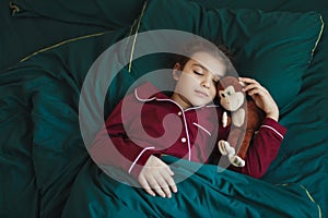 7-year-old girl in red pajamas sleeps on green bed with plush monkey toy. Baby looks peaceful and content as she sleeps.