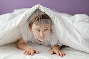 7 years old boy hiding in bed under a white blanket or coverlet