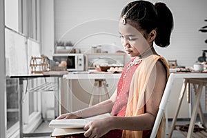 An 8-year-old Indian girl sitting in the kitchen reading a book and studying