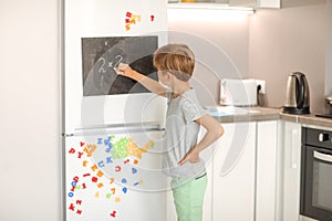 A 8-year-old schoolboy writes white chalk on a magnetic black board attached to a fridge