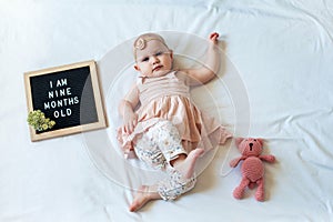 9 Nine months old baby girl laying down on white background with letter board and teddy bear. Flat lay composition.