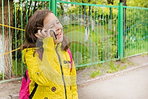 A 9-year-old girl in a yellow jacket speaks on a cell phone and smiles. The girl communicates with her mother on a mobile phone