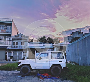 An abandon white jeep parked in a small park within a crowded residential housing area under pink yellow amber sky at dawn at dusk
