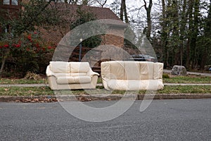 Abandoned beige couches on the grass by a curb in front of a house waiting to be removed by the garbage men