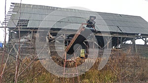 An abandoned board shed with a slate roof stands amidst a thicket of dry grass and bushes. A belt conveyor with an electric motor