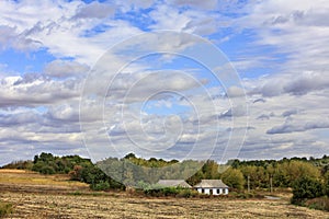 An abandoned rural house stands on the side of a rural road at the edge of a field under a cloudy sky