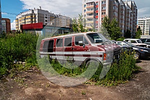 Abandoned rusting cars stand on the outskirts of a residential neighborhood, overgrown with grass against the backdrop of modern